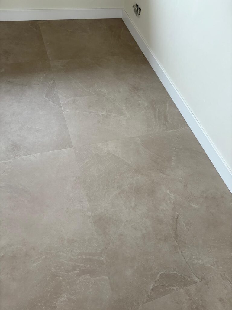 De vloer van een kamer met witte muren en een witte plint is bedekt met beige keramische tegels met een subtiel marmerpatroon. Aan de muur is een stopcontact zonder afdekplaatje te zien.