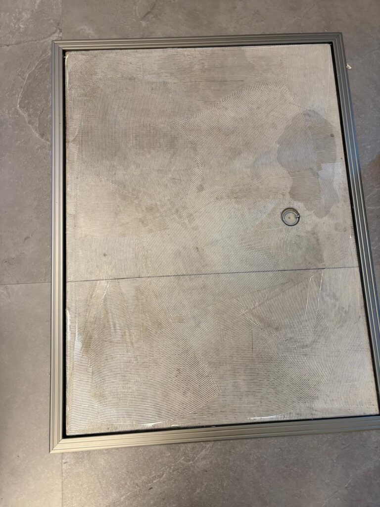 Een rechthoekig vloerluik met een metalen frame, ingebouwd in een grijze tegelvloer. Het luik vertoont zichtbare structuurpatronen en heeft rechtsboven een ronde handgreep.