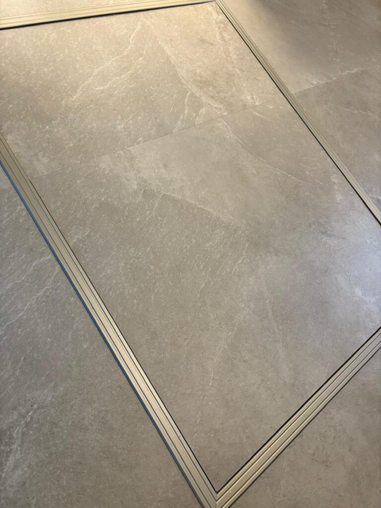 Grote beige vloertegels met een rechthoekig metalen inzetstuk, waardoor een omlijnd gedeelte in het tegeloppervlak ontstaat. De tegels hebben een subtiel natuursteenpatroon en een matte afwerking.